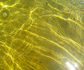 Sunlight refracting through transparent water and creates sun glare on golden sand of bottom. Top view of yellow ripple texture of liquid surface and sunny shining reflections.