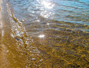 Transparent water and pebbles beach - close-up. Summer background image of sunlight reflecting and glistening on the water surface and wet small stones in foreground.