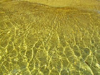 Transparent water surface with golden sun glare and sunny shining reflections on the sand. Top view of yellow ripple texture with sunlight refracting through liquid layer.