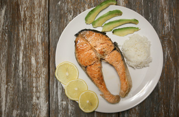 A piece of fry salmon fish steak on a white plate with spinach, rice and lemon, on  a gray wooden background. Omega 3 vitamin, healthy lifestyle. Natural vegetarian food. Top view. Copy space.