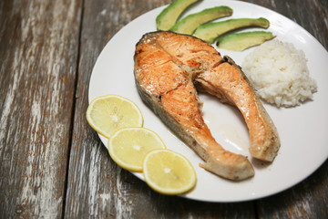 A piece of fry salmon fish steak on a white plate with spinach, rice and lemon, on  a gray wooden background. Omega 3 vitamin, healthy lifestyle. Natural vegetarian food. Top view. Copy space.