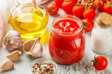Traditional tomato sauce in a glass jar with garlic and spices.  Ingredients and olive oil on the light surface