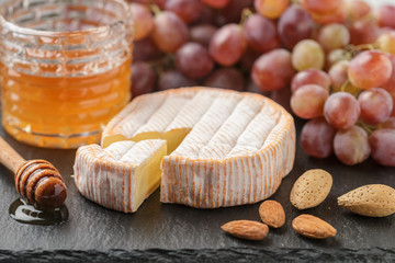 Delicatessen soft fragrant cheese with mold, honey, almonds and red grapes on a black slate plate....