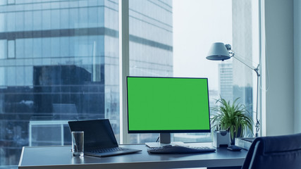 Shot of the Office Desk with Green Mock-up Screen Personal Computer Standing on it. Modern Stylish Room with a Big City Business District View.