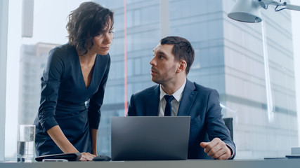 In the Office Female CEO Talks with Leading Businessman who sits at His Desk, Uses Laptop. They Discuss Ongoing Business Contracts and Deals. In the Background Big City View.