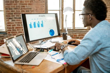 Young african man working with analitycs sitting at the beautiful working place with computer, laptop and documents