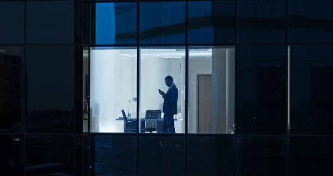 Aerial Shot: From Outside Into Office Building With Businessman Using Mobile Phone And Standing By The Office Window. Beautiful Shot Of The Financial Business District Skyscrapers. 