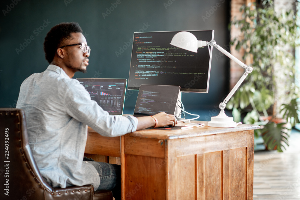 Wall mural young african male programmer writing program code sitting at the workplace with three monitors in t