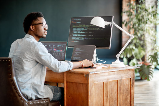 Young african male programmer writing program code sitting at the workplace with three monitors in the office. Image focused on the screen