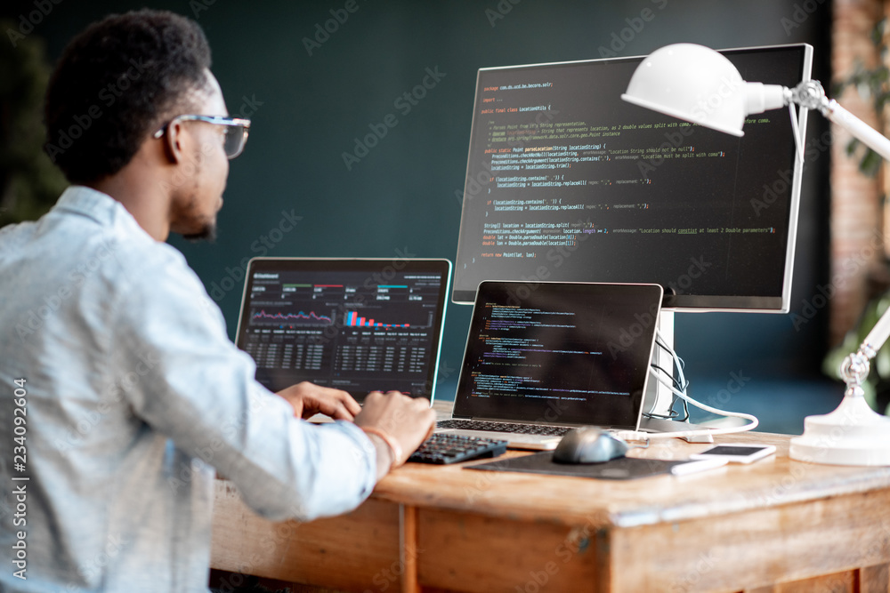 Wall mural young african male programmer writing program code sitting at the workplace with three monitors in t