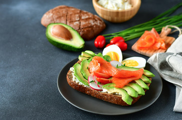 sandwich with avocado and salmon on a dark background, green onions and gluten-free grain bread, radishes and tomatoes, cottage cheese in a wooden plate. concept diet food, vegetarian, copy space