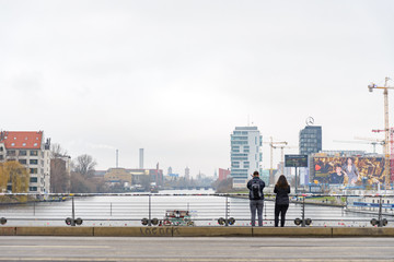 Fototapeta na wymiar Two tourists stand at Oberbaum bridge and look at scenery of river, east side gallery and riverside Spree river with cloudy gloomy sky in winter season. 