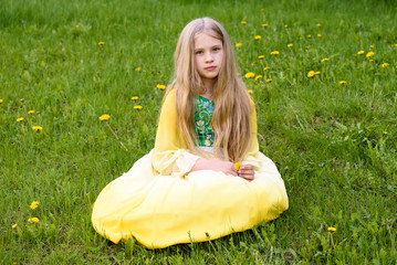 Blond young girl posing in a yellow green dress sitting on the grass with dandelions yellow flowers