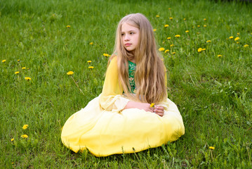 Blond young girl posing in a yellow green dress sitting on the grass with dandelions yellow flowers