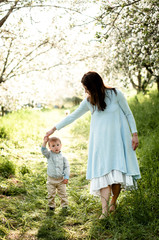 young beautiful mother in a blue lush dress holding a little son by the hand standing on a footpath in a blossoming apple orchard