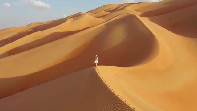 Young woman in a white summer dress and panama hat walking on the edge of a massive sand dune. Empty Quarter, Abu Dhabi, UAE.