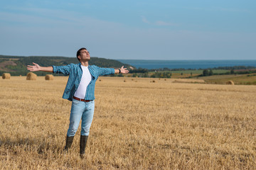 Naklejka na ściany i meble Young man in jeans, shirt, rubber boots in the field with his hands open, the concept of freedom, motivation, movement, front portrait