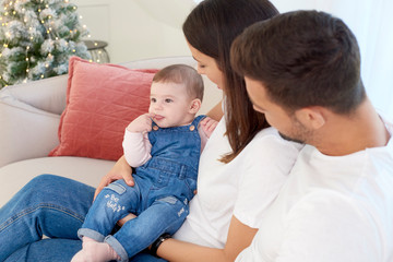 Obraz premium Happy young parents with their baby girl enjoy Christmas time while relaxing on the sofa