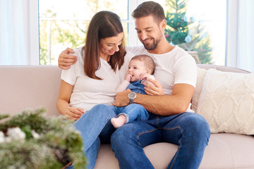 Happy young parents with their baby girl enjoy Christmas time while relaxing on the sofa