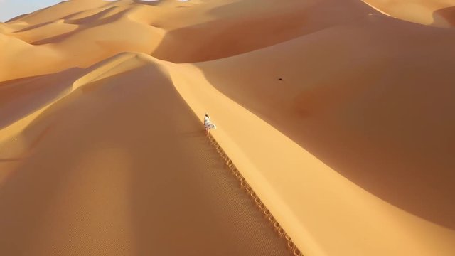 Aerial drone view zooming out of a young woman in white summer dress and hat walking up massive yellow and gold sand dunes with shadows in empty quarter during sunset.