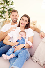 Obraz premium Happy young parents with their baby girl enjoy Christmas time while relaxing on the sofa