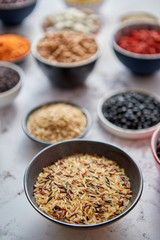 Raw wold rice seeds in ceramic bowl. Composition of superfoods in background. Placed on white rusty table. Selective focus.