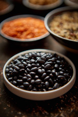 Raw black beans in ceramic bowl. Composition of superfoods in background. Placed on dark rusty table. Selective focus.