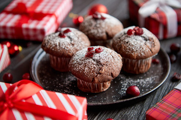 Christmas chocolate delicious muffins served on black ceramic plate. Sprinkled with powder sugar. Cranberries on top. Xmas gifts and decorations on sides.