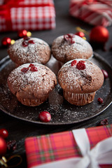 Christmas chocolate delicious muffins served on black ceramic plate. Sprinkled with powder sugar. Cranberries on top. Xmas gifts and decorations on sides.