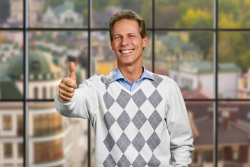 Joyful mature man giving thumb up sign. Cheerful caucasian man showing thumb up on abstract window background. Human gestures and emotions.