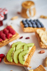 Assortment of healthy fresh breakfast toasts. Bread slices with peanut butter and various fruits and ingredients on side. Placed on white wooden table. Top view, with copy space. Selective focus