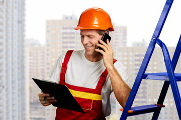 Happy builder talking on phone. Smiling engineer talking on smartphone while holding clipboard with documents. Discussing new project.