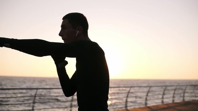 Full lenght of a young athlete in black shirt and red sneakers while training process on the promenade in front the sea in the early morning. Man boxing with invisible opponent, punching - wearing