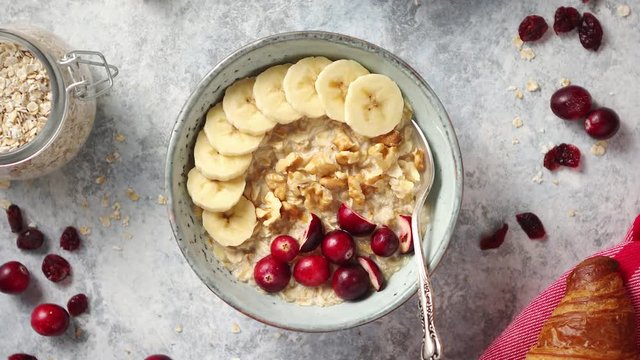 Ceramic bowl of oatmeal porridge with banana, fresh cranberries and walnuts on stone table top view in flat lay style. Healthy breakfast and diet food.