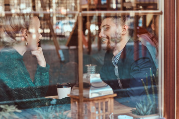 Attractive young couple looking at each other and talking while sitting in the restaurant behind the window.