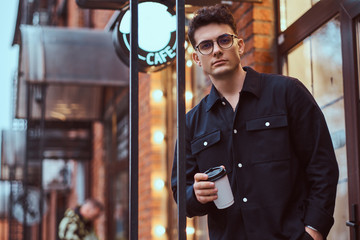A young handsome man holding a takeaway coffee while standing near a cafe outside.