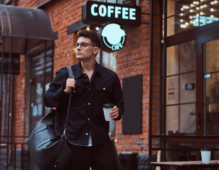 A young handsome man holding a takeaway coffee while standing near a cafe outdoors.