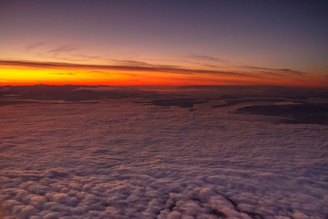 The golden sun casts its rays across the tops of clouds at sunset while lit cities await below