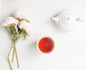 Directly above view of pale pink peonies, tea in cup and teapot on white background (selective focus)