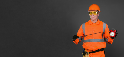 Happy builder worker is showing on a red alarm clock in his hand isolated on gray background with copy space. Time to work. Building construction time.