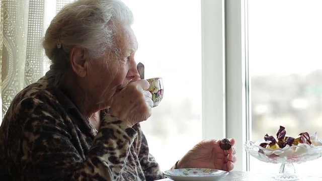 Old woman 90 years old looking out the window and drinking tea or coffee