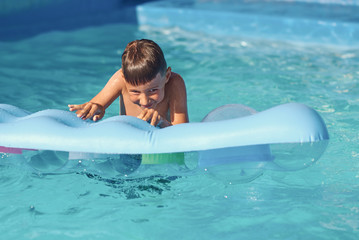 Happy Smiling Caucasian boy trying to get out from water on inflatable mattress in swimming pool at...