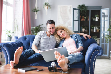 Couple searching locations a trip with laptop sitting on a couch in the living room at home.