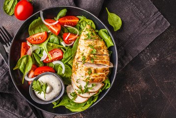 Grilled chicken breast salad with spinach, tomatoes and Caesar dressing, dark background.