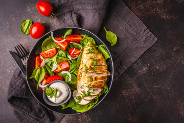 Grilled chicken breast salad with spinach, tomatoes and Caesar dressing, dark background.