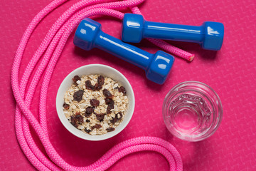 healthy lifestyle concept, on the fitness mat there is a bowl with muesli, two dumbbells, a jump rope and a glass of water