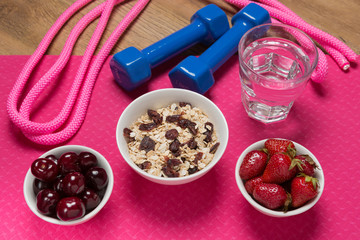 the concept of sports and healthy eating, on the mat for fitness is three bowls of fruit and cereal, two dumbbells and a glass of water