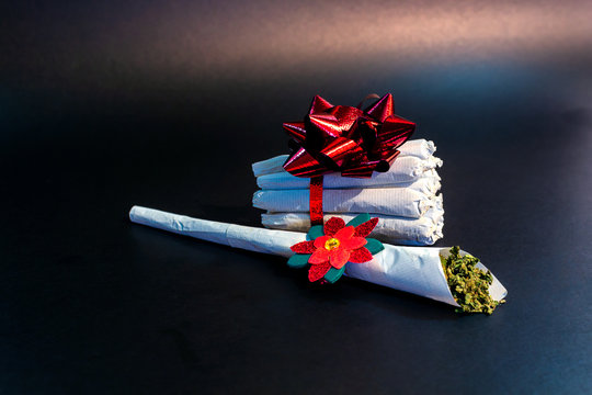 A large marijuana joint with a festive decoration on it in front of a bundle of joints tied in ribbon and a bow isolated.