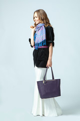 Stylish beautiful woman wearing a scarf around her neck holding a fashionable handbag in her hand. Studio photography on a blue background. Vertical orientation.
