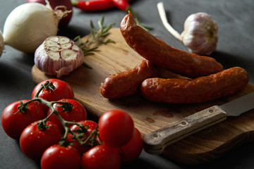 three delicious sausages with different vegetables on a wooden board, knife on a black table